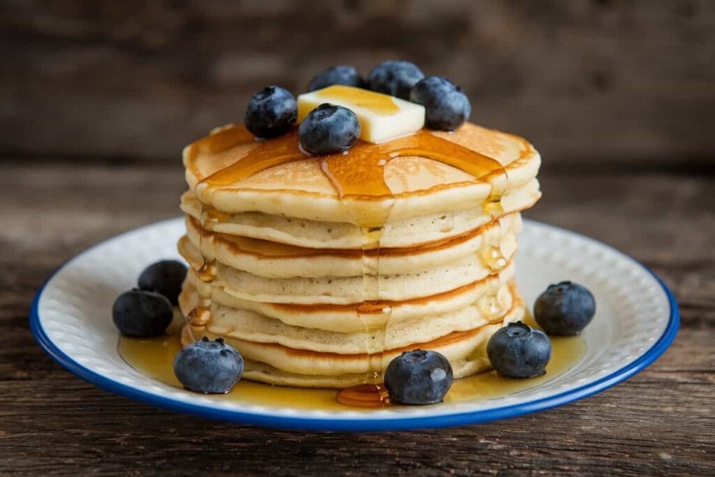 A stack of fluffy pancakes with syrup, blueberries, and butter on a plate.